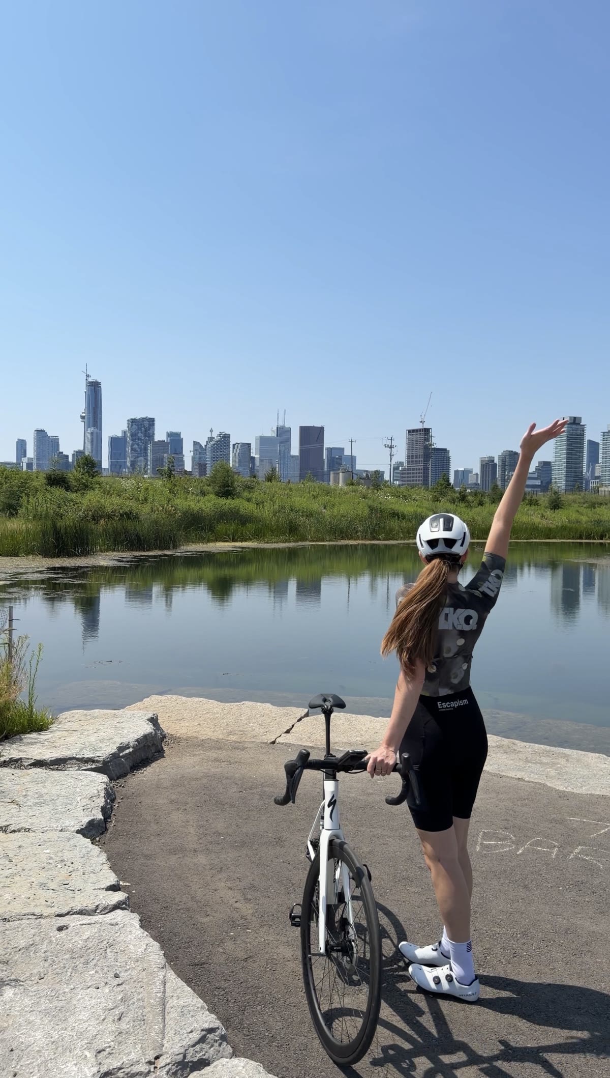 Bailey standing with her bike at Biidaasige Park, waving toward the Toronto skyline across the water