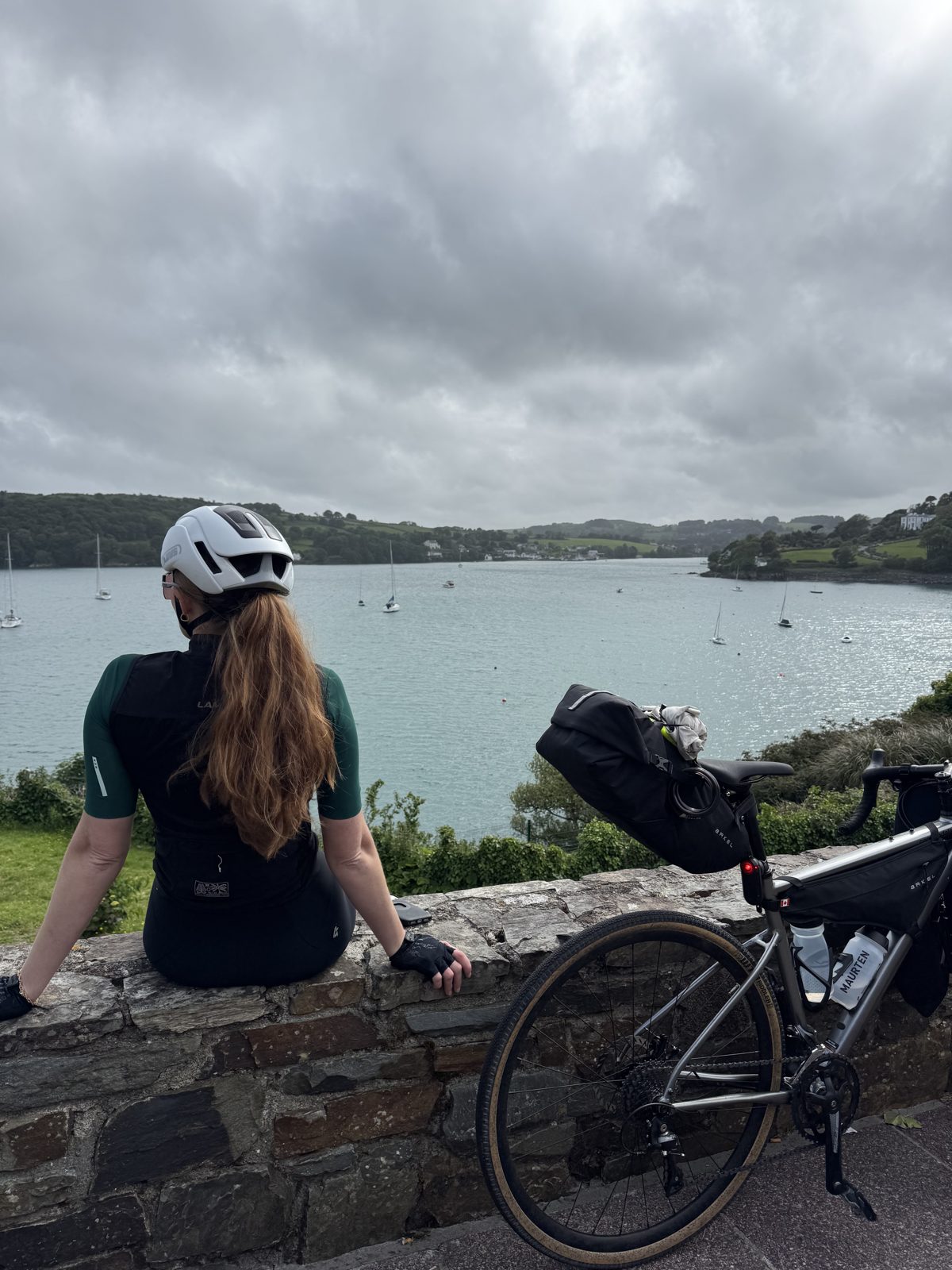 Bailey resting on a stone wall beside the loaded bike, looking out over a West Cork harbour dotted with sailboats