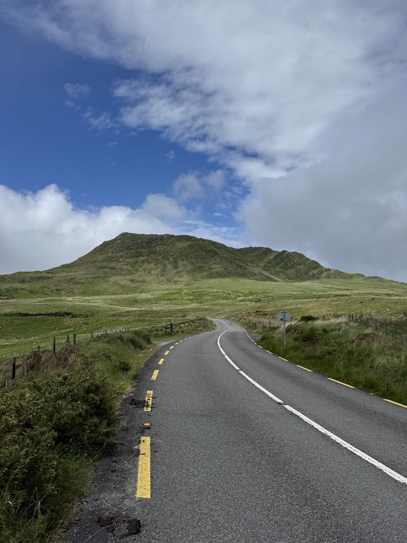 An empty road climbing toward a green Kerry mountain under a big blue sky with broken cloud
