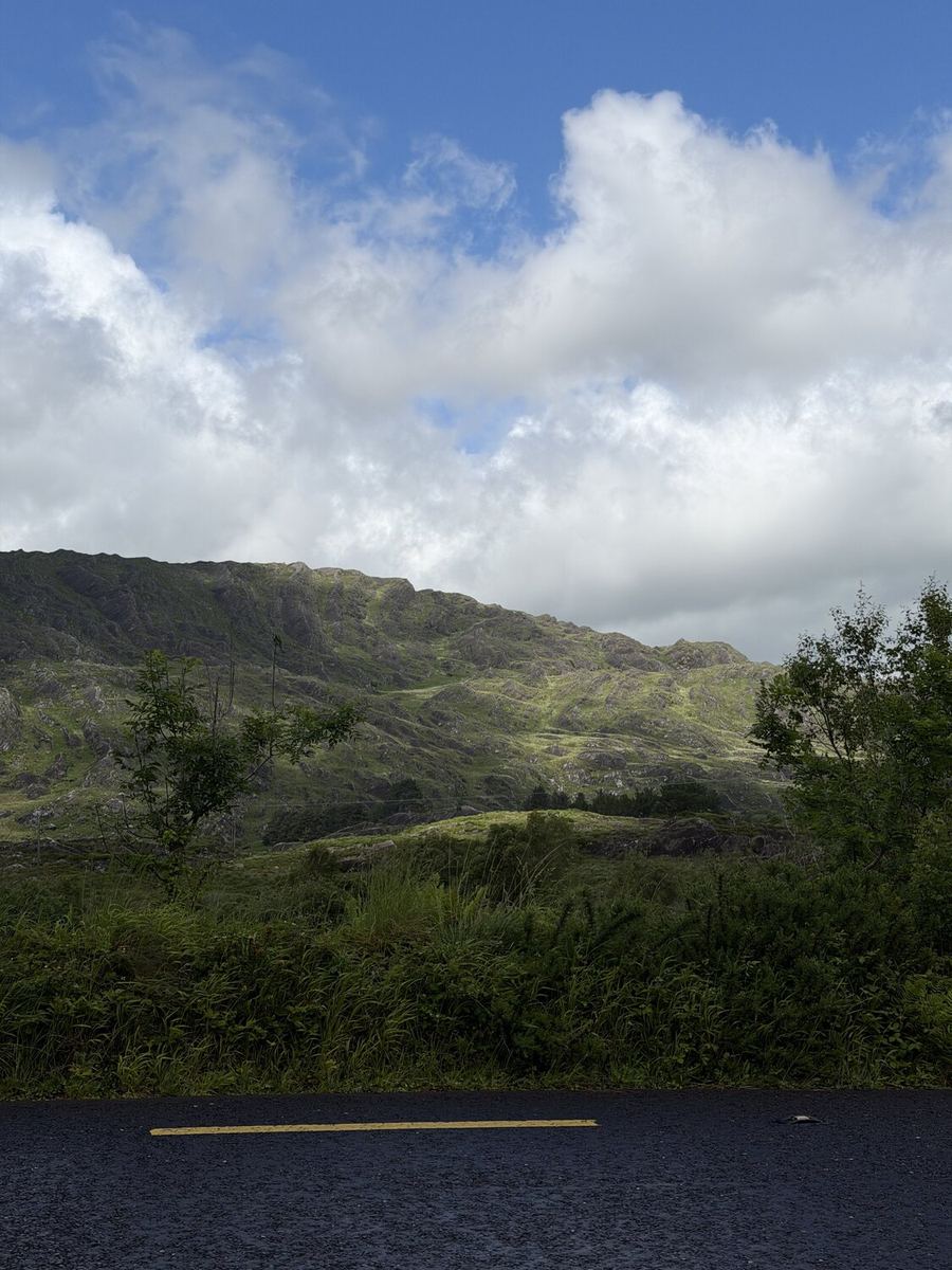 A rugged green mountainside rising above the road on the Beara, clouds breaking to blue overhead