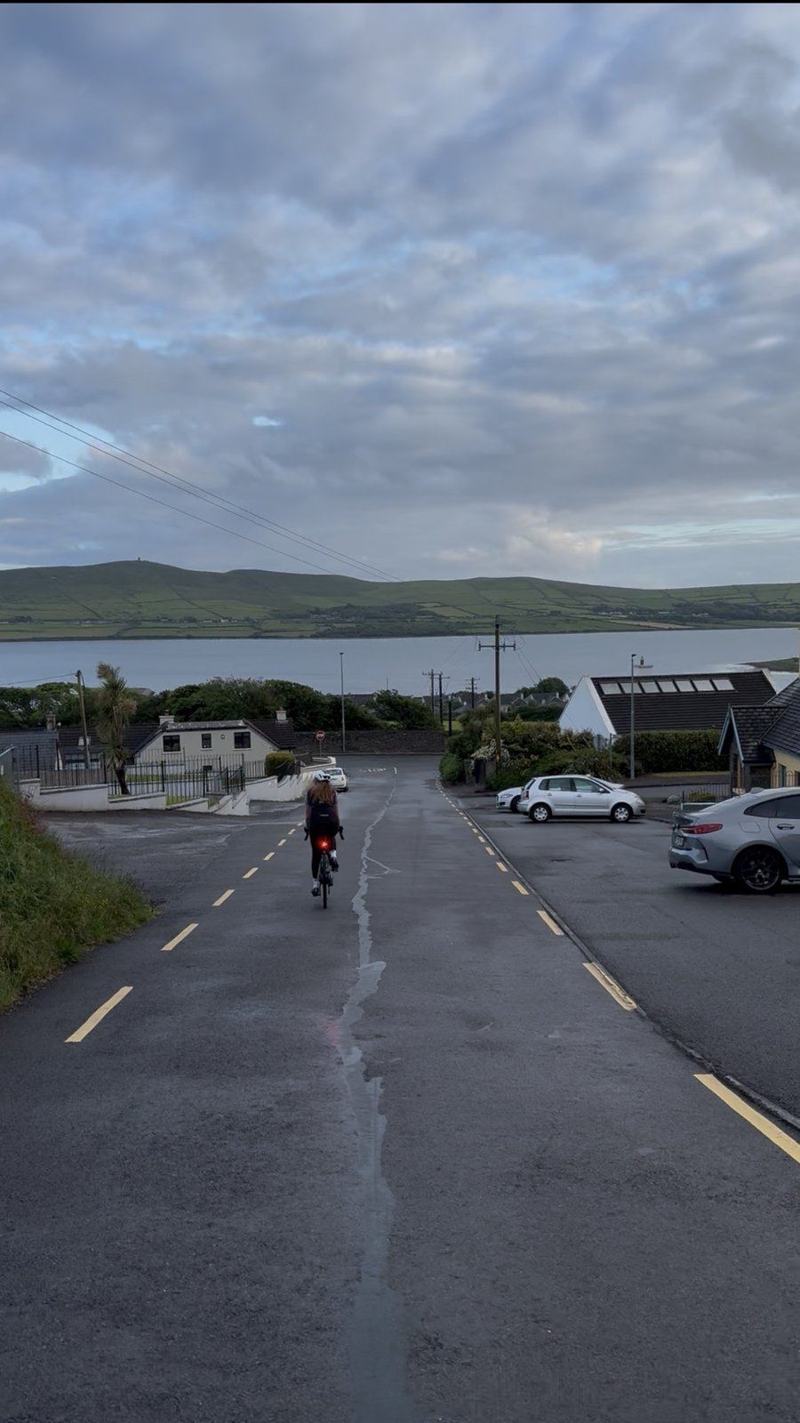 A rider freewheeling down a road toward Ventry harbour with the hills of the Dingle Peninsula across the bay